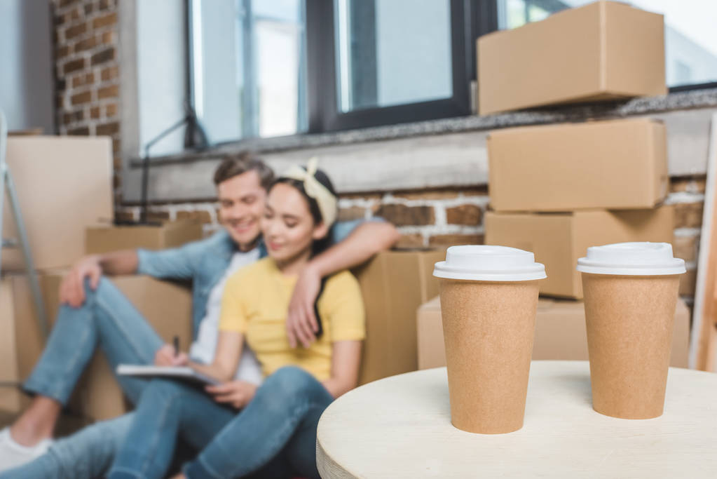 beautiful young couple sitting on floor while moving into new home with coffee to go on foreground - Photo, Image