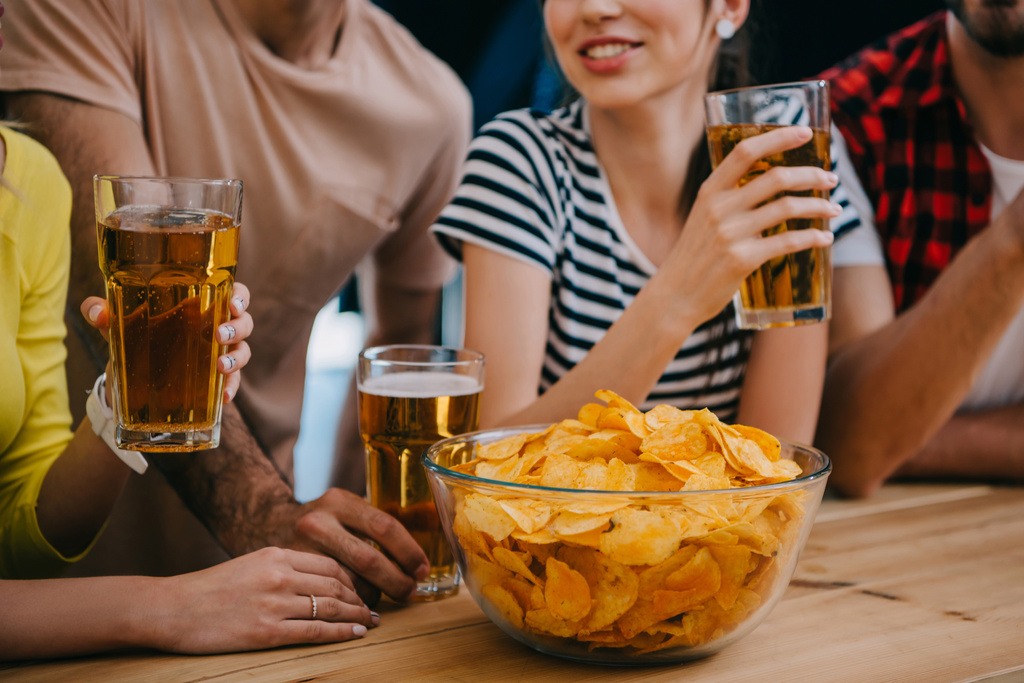 tiro recortado de grupo de amigos com cerveja e tigela de batatas fritas sentado no balcão de bar durante relógio de jogo de futebol
 - Foto, Imagem