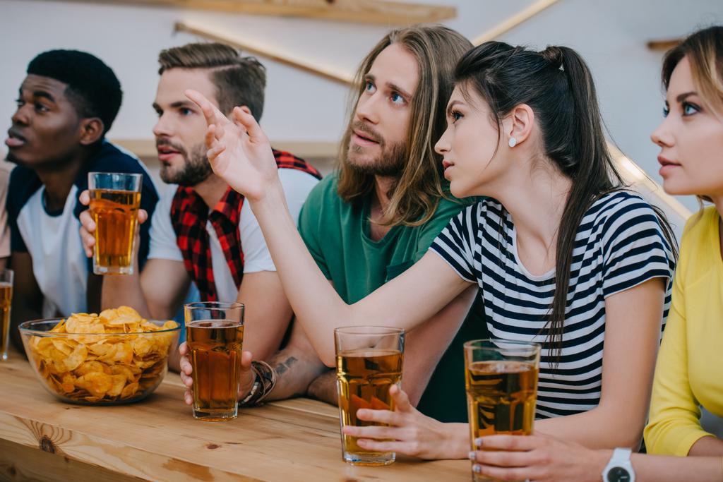 fiatal nő barátja ujjal mutat, és a gazdaság sör üveg, míg a többi meg nézi labdarúgó mérkőzés a bar  - Fotó, kép
