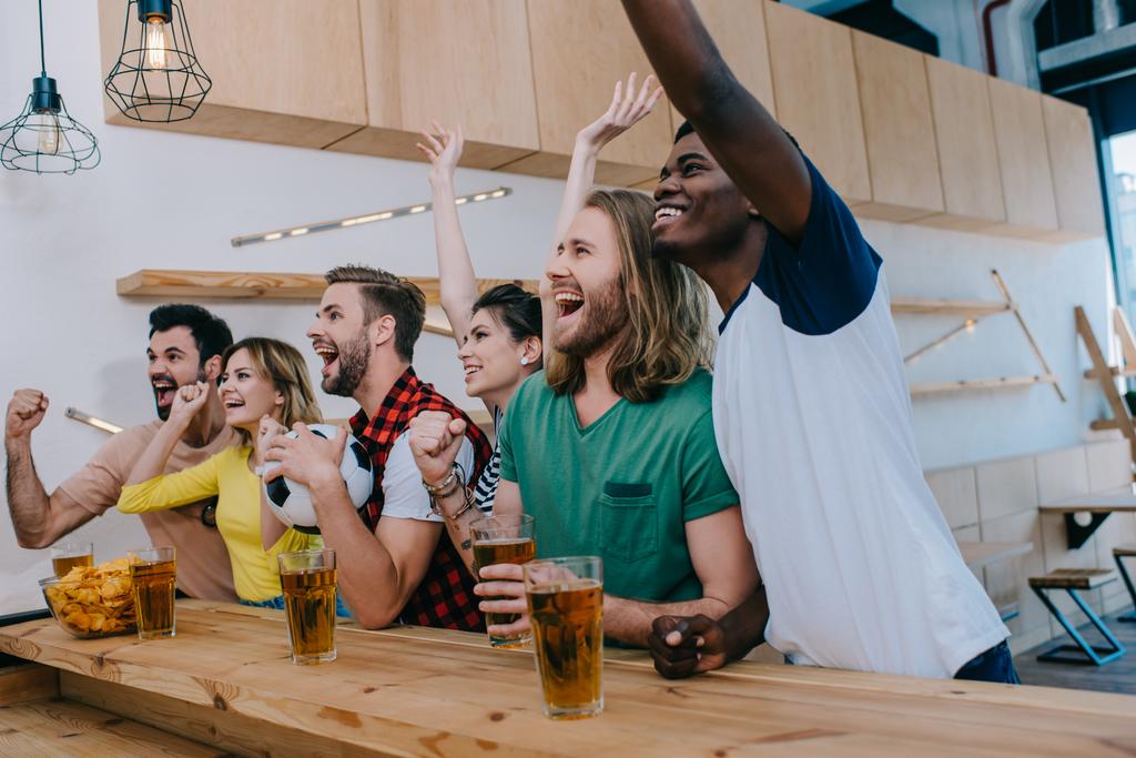 взволнованные мультикультурные друзья празднуют, жестикулируют руками и смотрят футбольный матч в баре
  - Фото, изображение