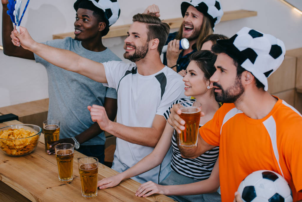 glimlachend multiculturele vrienden in voetbal hoeden vieren, drinken bier en kijken naar de voetbalwedstrijd op balk  - Foto, afbeelding