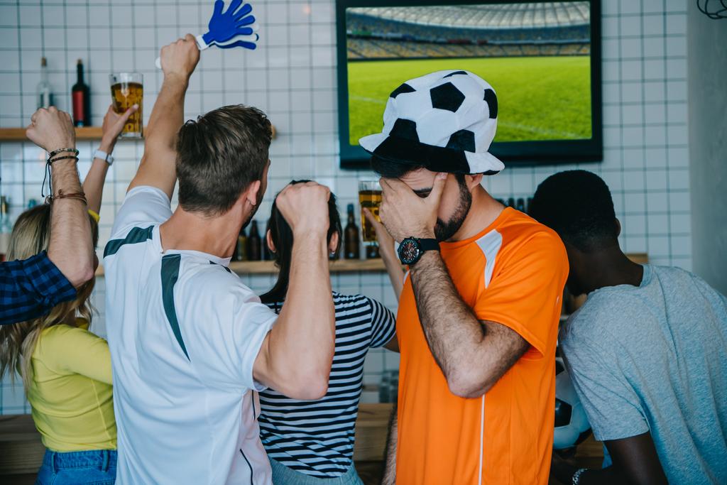 gefrustreerd jongeman in Voetbal bal hoed fan t-shirt bedrijf bekleding ogen met de hand palm terwijl zijn vrienden vieren en doen ja gebaren tijdens horloge van soccer match op tv-scherm op balk  - Foto, afbeelding