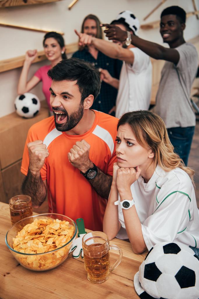 емоційні мультикультурні шанувальники футболу, що вказують пальцями і дивляться футбольний матч в барі з чіпсами і пивними окулярами
  - Фото, зображення