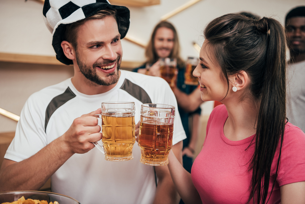 junger glücklicher Mann mit Fußballmütze, der mit Freundin Biergläser klirrt, während ihre multikulturellen Freunde in der Bar Fußballspiele gucken  - Foto, Bild