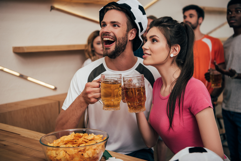 uomo eccitato in cappello da pallone da calcio clinking bicchieri di birra con la ragazza mentre i loro amici multiculturali guardando la partita di calcio al bar
  - Foto, immagini