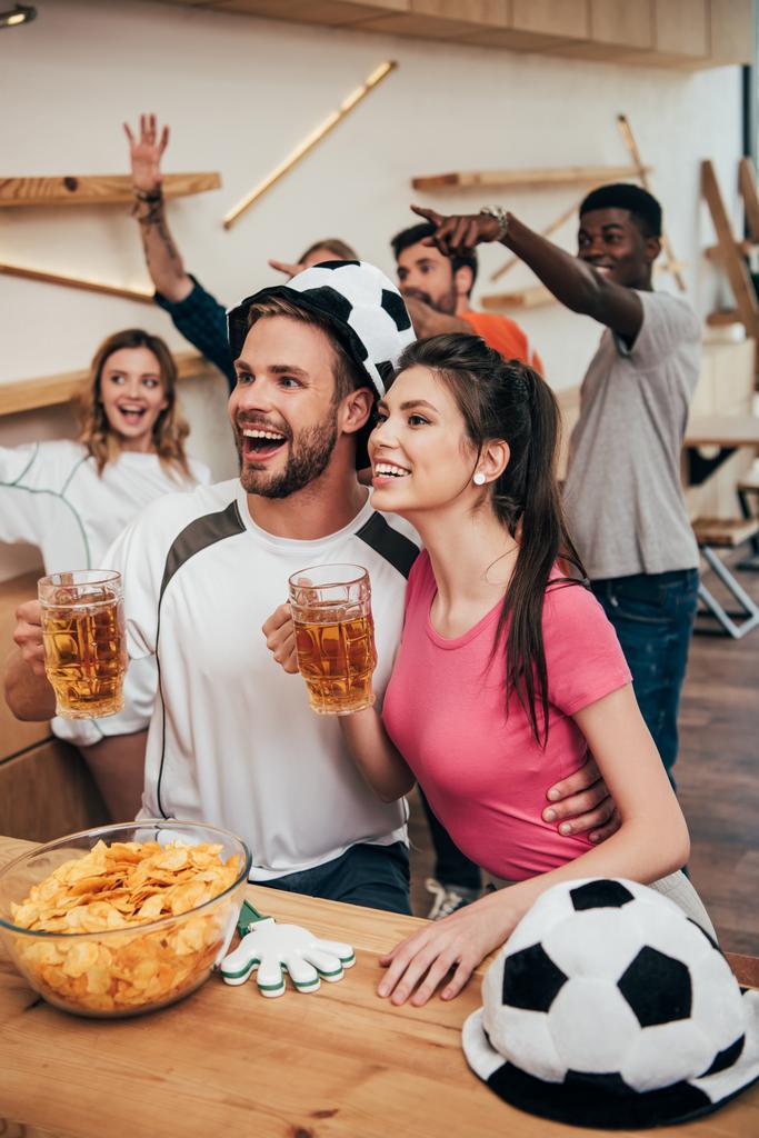 homme heureux dans le chapeau de ballon de football embrassant petite amie et tenant de la bière tandis que leurs amis célébrant derrière et gesticulant par les mains pendant la montre du match de football au bar
  - Photo, image