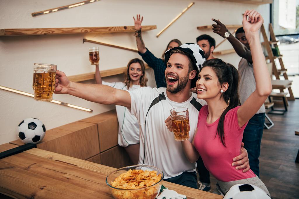 vista de ángulo alto del hombre feliz en sombrero de fútbol abrazando novia y sosteniendo la cerveza mientras sus amigos celebran detrás y viendo el partido de fútbol en el bar
  - Foto, imagen