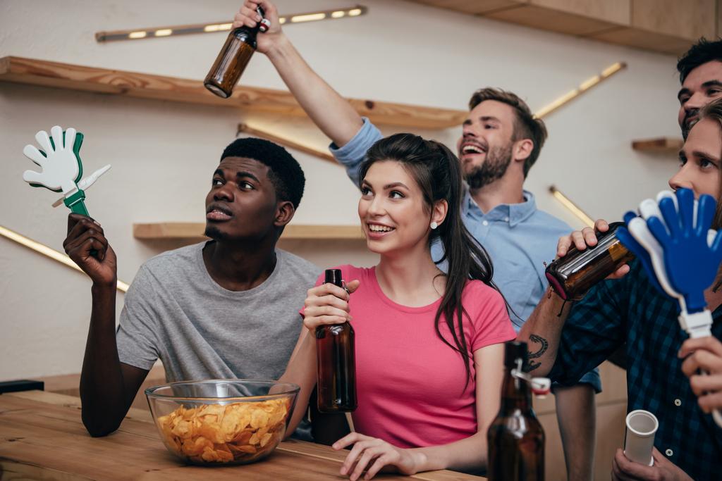 щасливі багатокультурні друзі, що святкують з пляшками пива та м'ячиками під час спостереження за футбольним матчем у барі
  - Фото, зображення