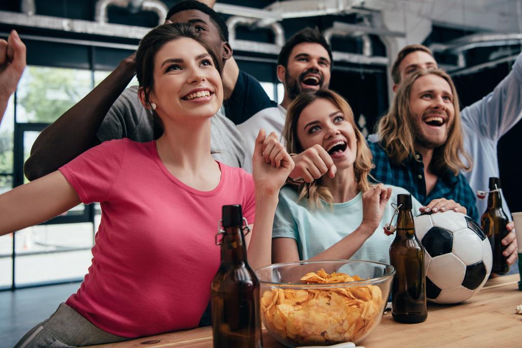 щаслива багатоетнічна група друзів, які святкують і дивляться футбольний матч в барі з чіпсами, пивом і футбольним м'ячем
  - Фото, зображення