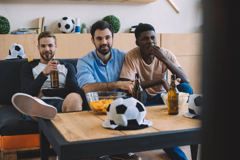 jonge multiculturele vrienden kijken naar voetbal wedstrijd in de buurt van tabel met chips, popcorn en bal hoeden thuis - Foto, afbeelding