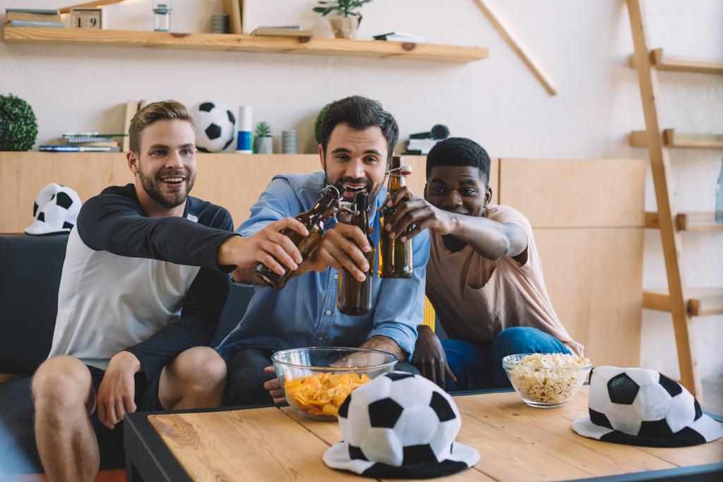 Lächelnde junge multikulturelle Freunde klappern mit Bierflaschen und feiern während des Fußballspiels zu Hause - Foto, Bild
