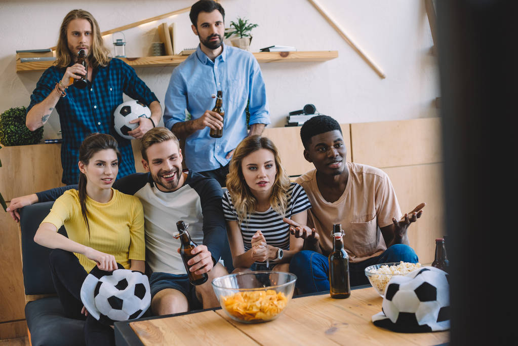 sorrindo amigos multiculturais garrafas de cerveja sentado no sofá perto da mesa com pipocas e batatas fritas assistindo jogo de futebol em casa
 - Foto, Imagem