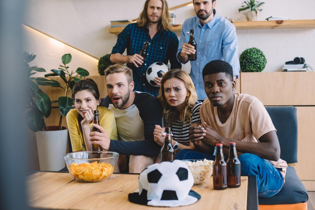 multikulturní přátel pivní lahve, sedí na pohovce u stolu s popcorn a hranolky, sledovat fotbalový zápas doma - Fotografie, Obrázek