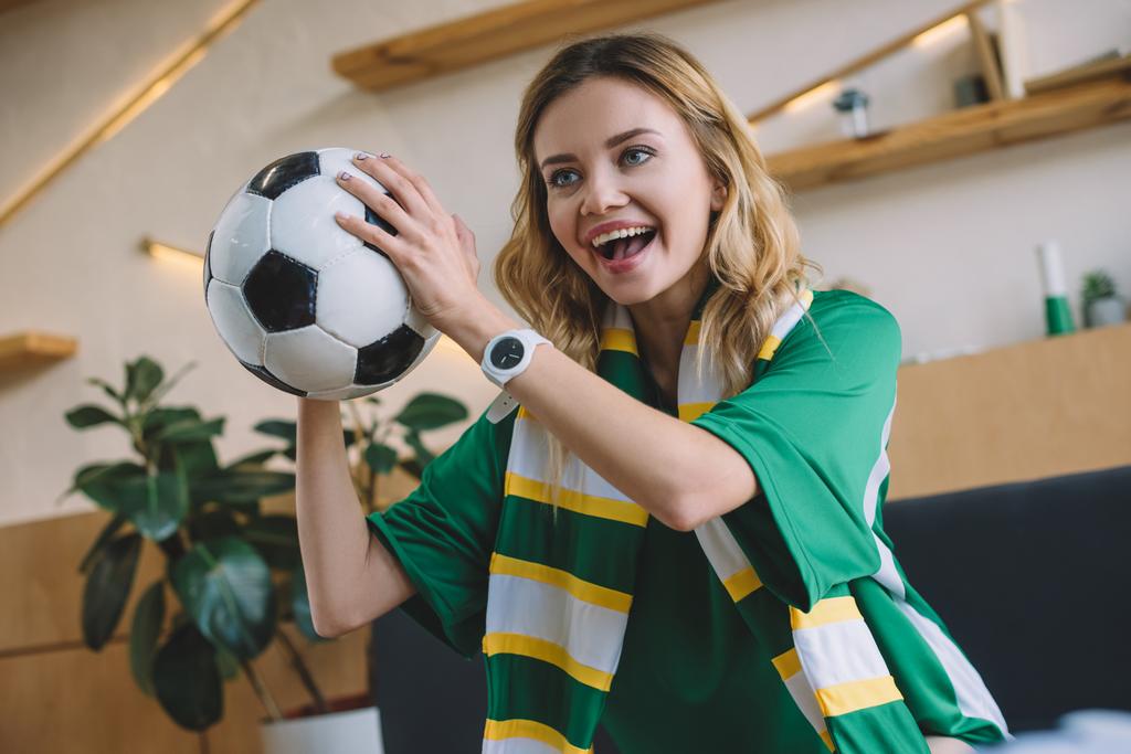 joven entusiasta del fútbol femenino en camiseta verde y bufanda sosteniendo la pelota y celebrando durante el reloj del partido de fútbol en casa
 - Foto, imagen
