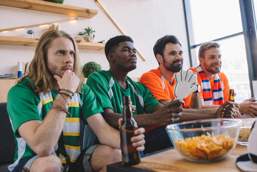 grupo de amigos masculinos multiculturales en verde y naranja ventilador camisetas viendo partido de fútbol cerca de la mesa con patatas fritas y palomitas de maíz en casa
 - Foto, Imagen