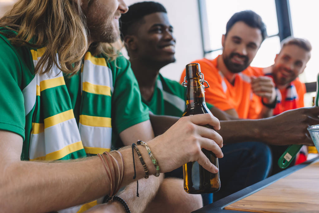 przycięte strzał człowieka w green fan Koszulka i szalik, trzymając butelkę piwa podczas jego wielokulturowy mężczyzna przyjaciół oglądanie piłka nożna mecz w domu - Zdjęcie, obraz