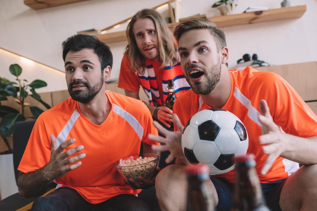 emotionele jonge mannelijke voetbalfans in oranje t-shirts met bal en popcorn kijken naar voetbalwedstrijd thuis - Foto, afbeelding