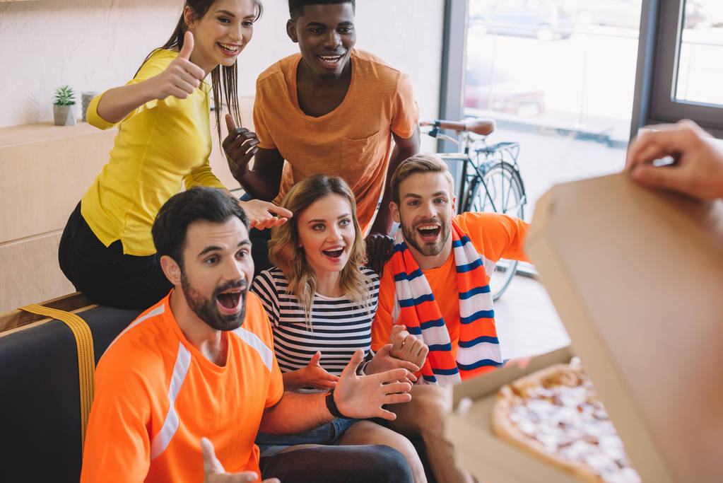 recortado tiro de la persona sosteniendo pizza y grupo excitado de amigos multiculturales en casa
 - Foto, Imagen