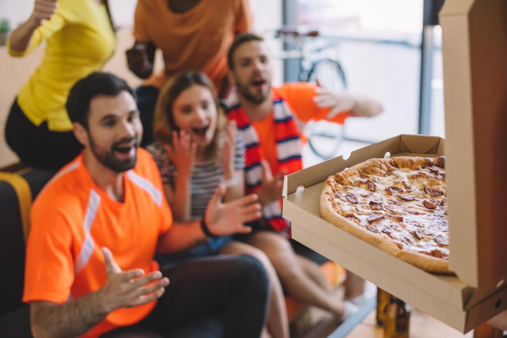 close up shot of pizza in box and excited group of friend sitting on sofa at home - Photo, Image
