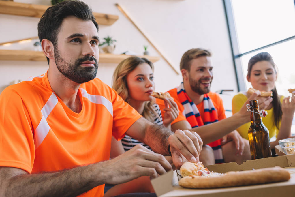 Nuori mies ottaa pizzaa laatikosta, kun hänen ystävänsä syövät pizzaa ja katsovat jalkapalloa kotona
 - Valokuva, kuva