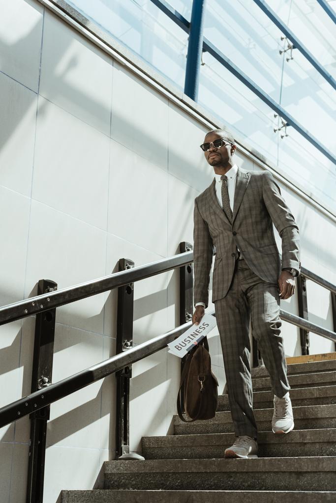Hombre de negocios afroamericano con traje sosteniendo periódico y maletín mientras camina por las escaleras
 - Foto, Imagen