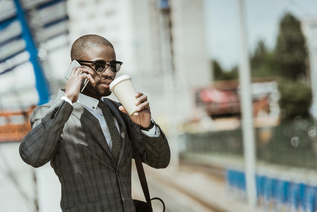 スマート フォンとコーヒー カップが列車を待っているとスーツを着てアフリカ系アメリカ人のビジネスマン - 写真・画像