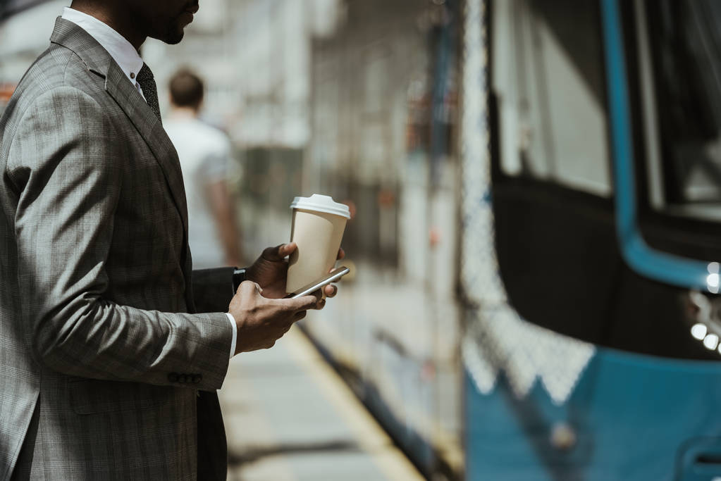 Vista recortada del joven hombre de negocios afroamericano con teléfono inteligente y taza de café tomando el tren
 - Foto, Imagen