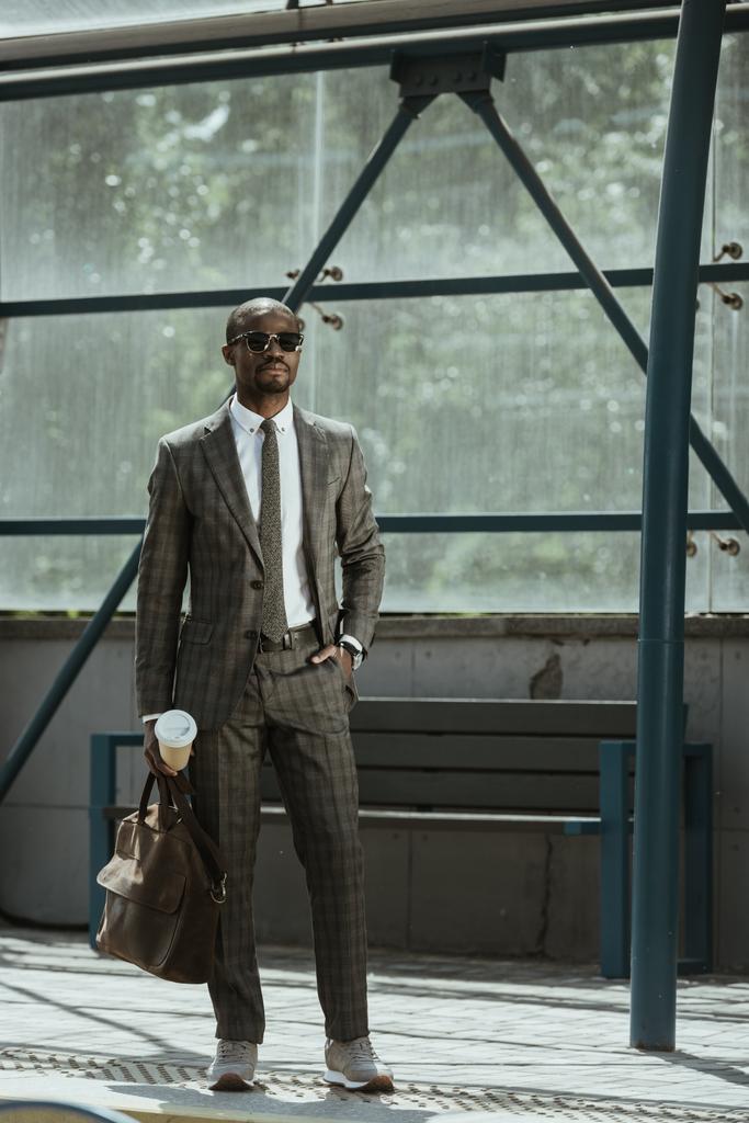 Hombre de negocios afroamericano con traje con maletín y taza de café de pie en la estación de transporte público
 - Foto, Imagen