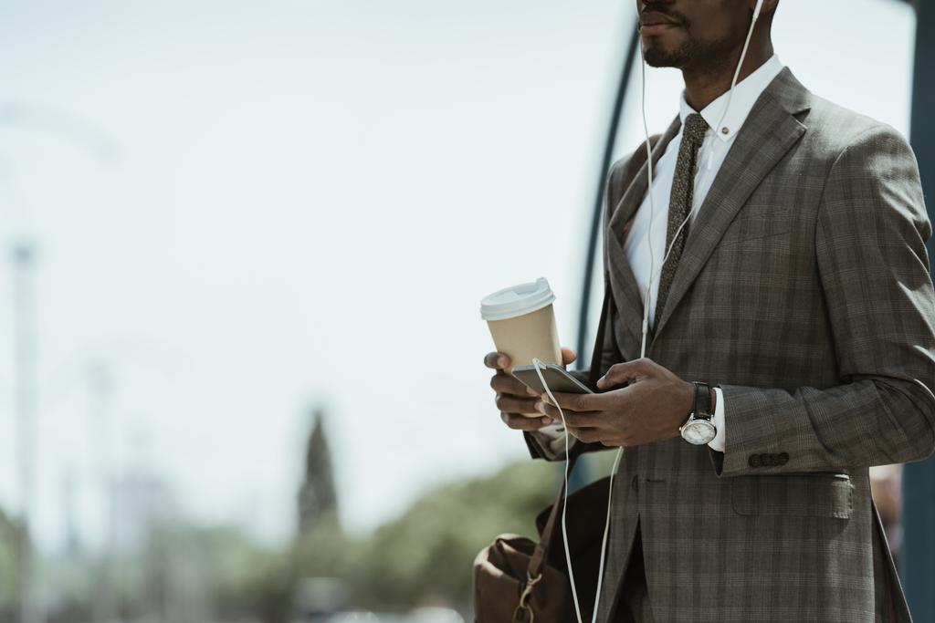 Ausgeschnittene Ansicht eines afrikanisch-amerikanischen Geschäftsmannes im Anzug, der Musik hört und eine Kaffeetasse auf dem Bahnhof hält - Foto, Bild