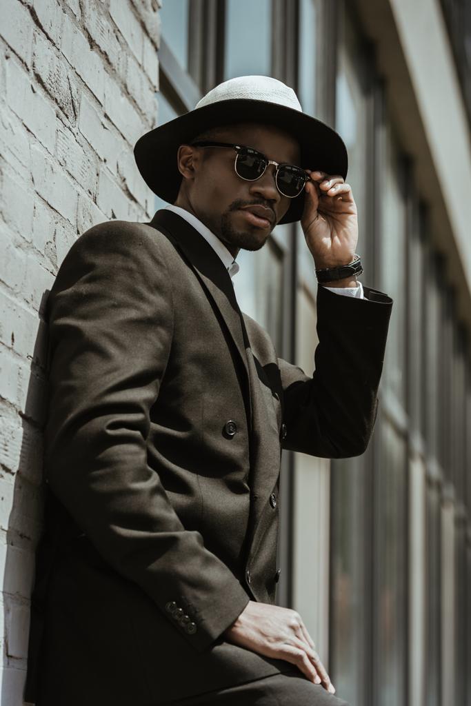 Elegante empresário afro-americano vestindo chapéu fedora e óculos de sol posando por parede de construção
  - Foto, Imagem