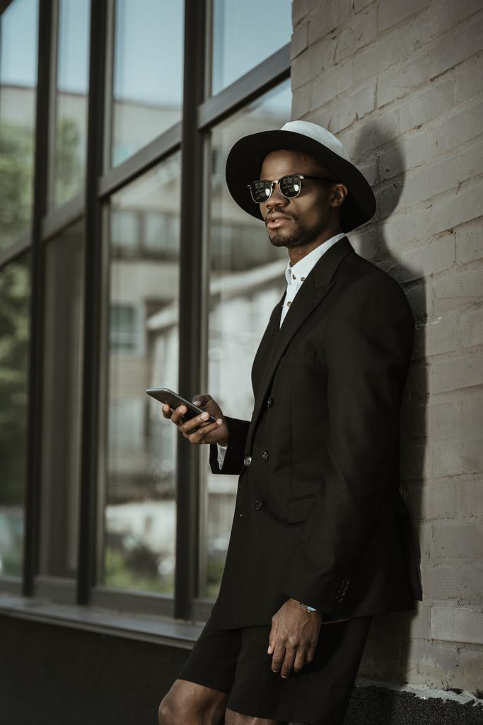 Muodikas afrikkalainen amerikkalainen mies yllään fedora hattu ja käyttämällä älypuhelinta kaupungissa
 - Valokuva, kuva