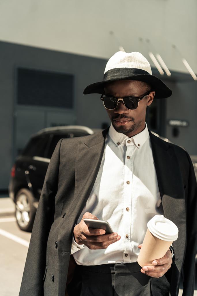 Beau jeune homme afro-américain utilisant un smartphone et tenant une tasse de café
 - Photo, image