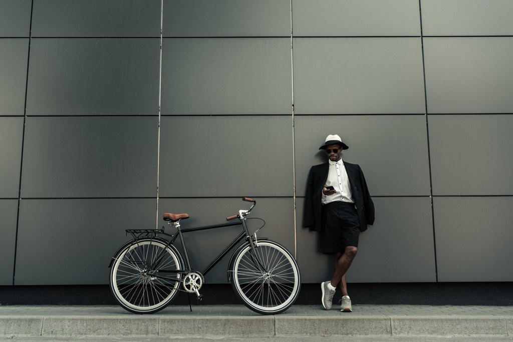 Модный африканский американец в шляпе-шляпе с помощью смартфона, стоящий у велосипеда
 - Фото, изображение
