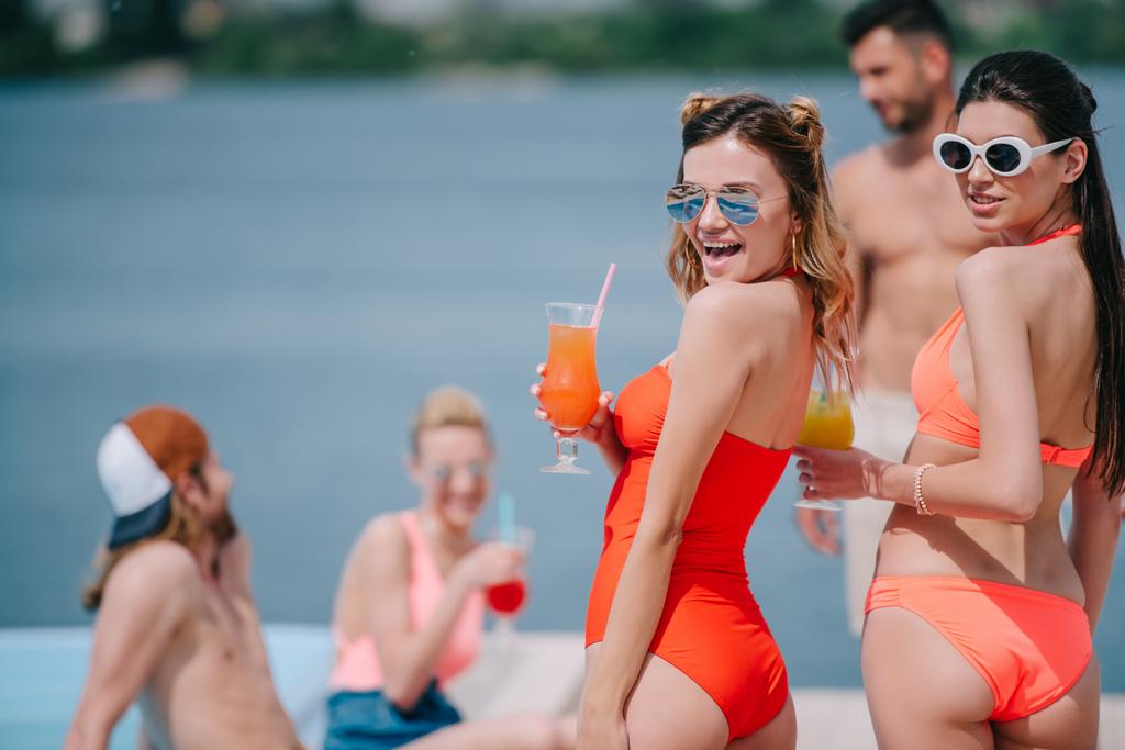красивые девушки в солнечных очках и купальниках, держащие коктейли и улыбающиеся перед камерой на пляже
 - Фото, изображение