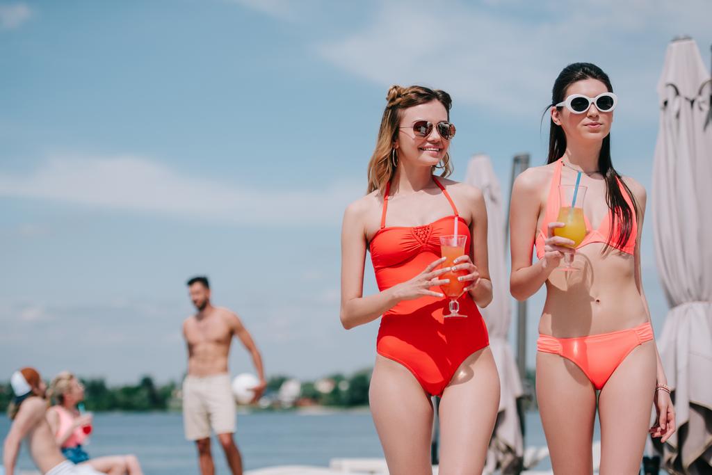 beautiful young women in sunglasses and swimwear holding glasses with cocktails at beach - Photo, Image