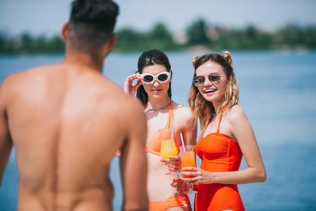 selective focus of girls in swimwear holding cocktails and looking at young man on beach - Photo, Image