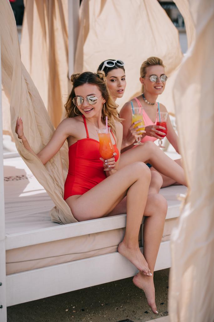 happy girlfriends in swimwear and sunglasses drinking cocktails in bungalow on beach  - Photo, Image