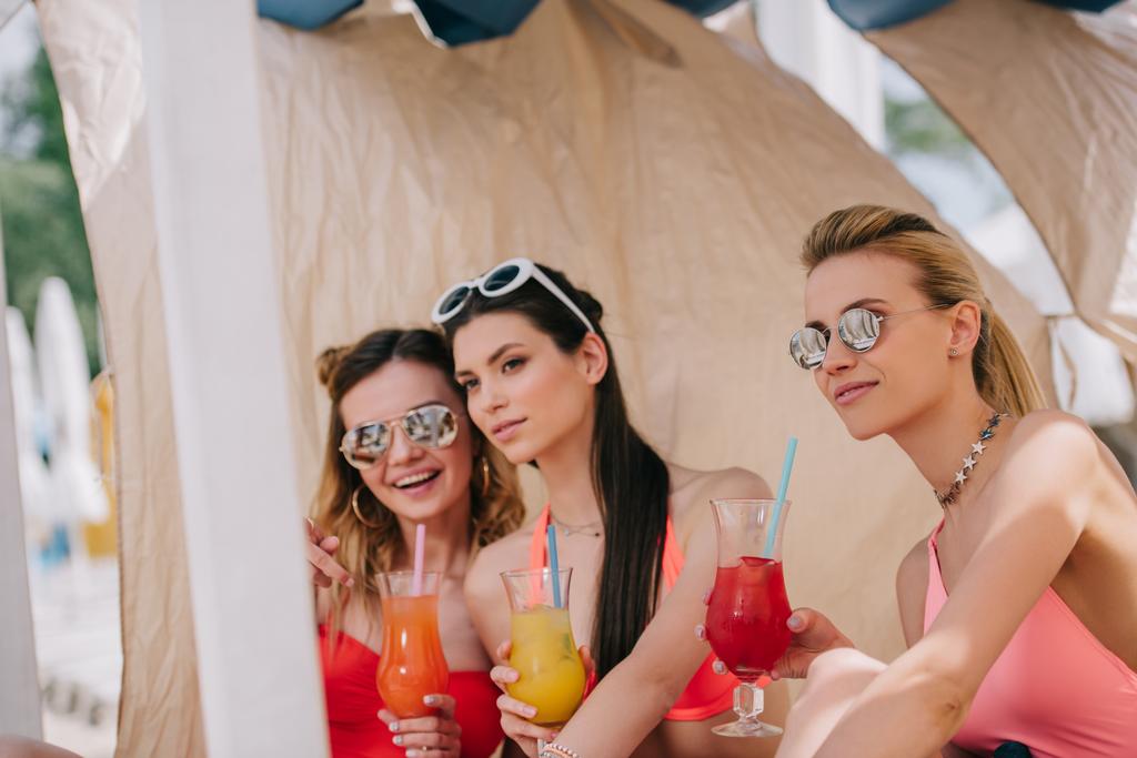красивые молодые женщины в купальниках и солнцезащитных очках, пьющие коктейли в бунгало на пляже
  - Фото, изображение