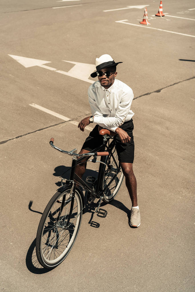 Модный африканский американец в шляпе-шляпе и солнцезащитных очках, позирующий на городском велосипеде
 - Фото, изображение