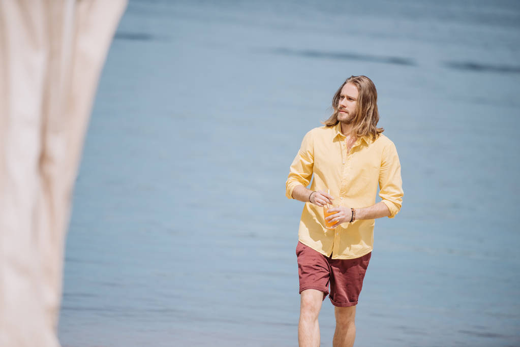 handsome young man holding glass with cocktail and looking away while walking at beach   - Photo, Image
