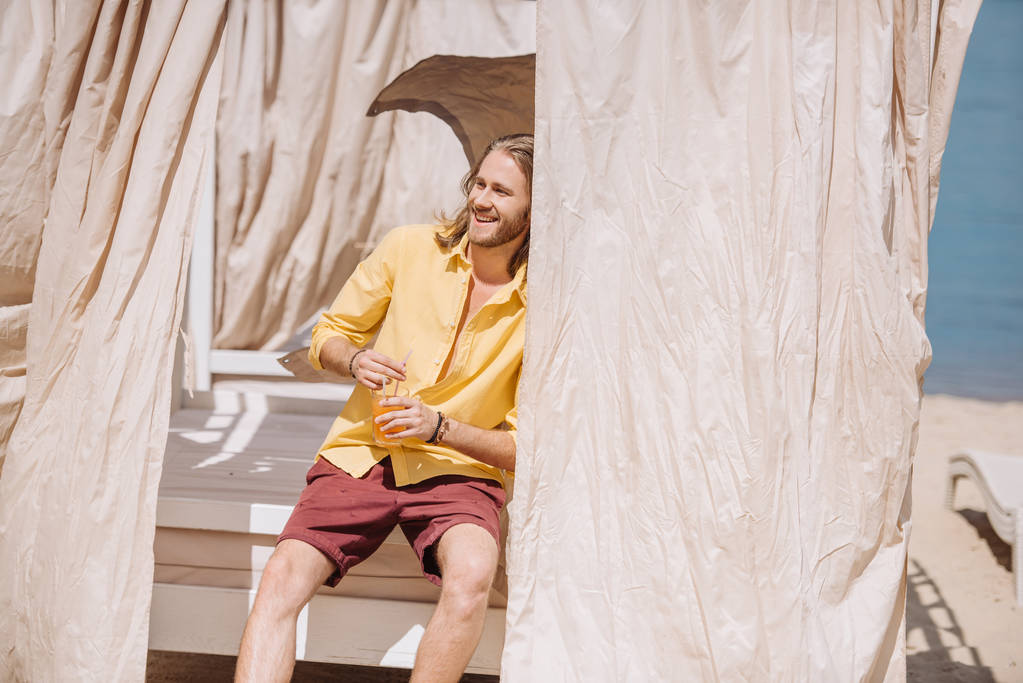 szép, mosolygós fiatalember gazdaság pohár koktél ülve Beach bungalow - Fotó, kép