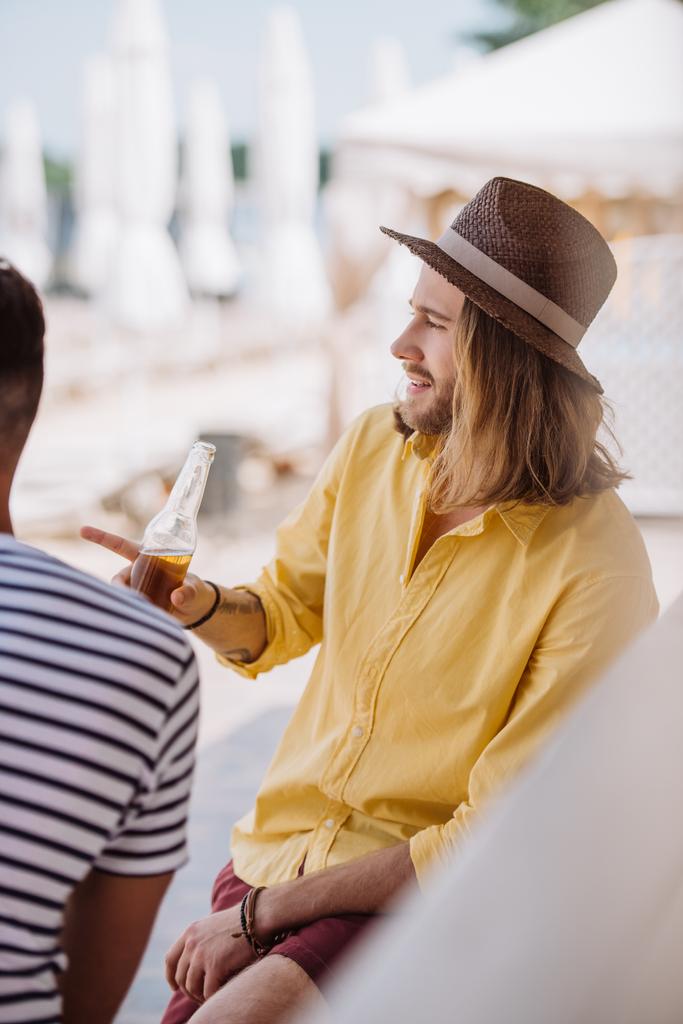 junger Mann hält Bierflasche in der Hand und zeigt weg, während er mit Freund an der Strandbar sitzt - Foto, Bild