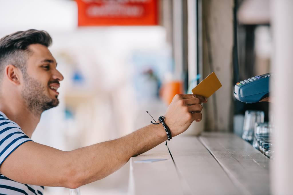 вид сбоку улыбающегося молодого человека, расплачивающегося кредитной картой в пляжном баре
 - Фото, изображение