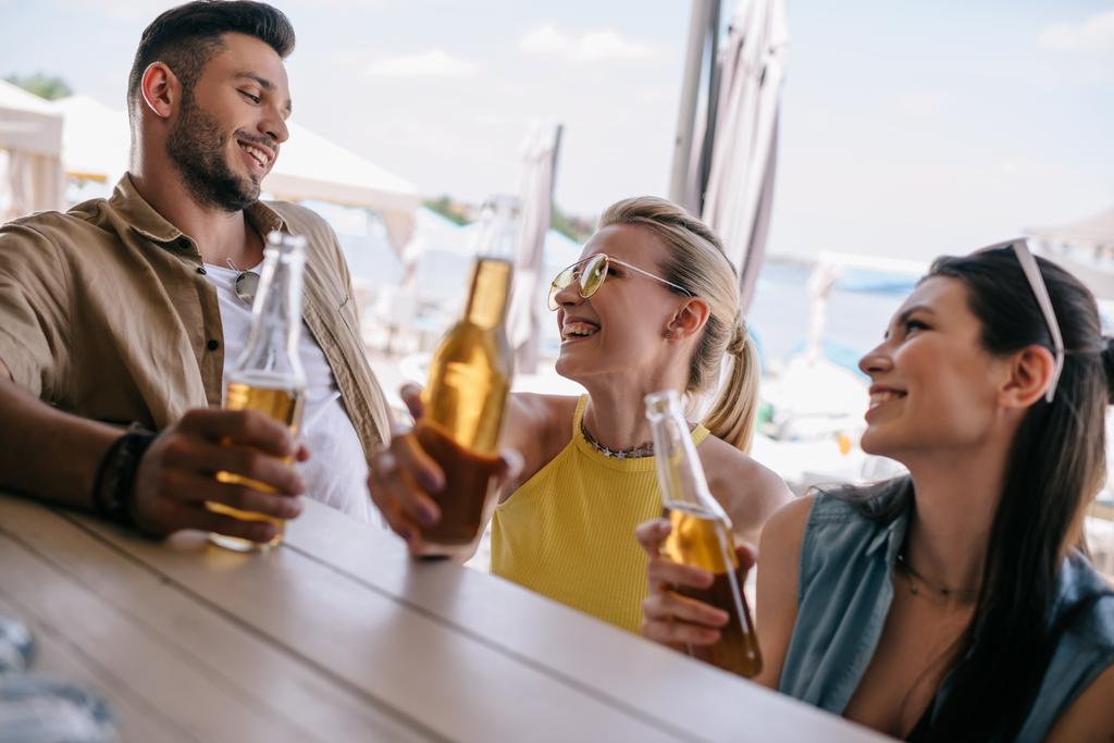 happy young man looking at beautiful smiling girls and drinking beer together at beach bar - Photo, Image
