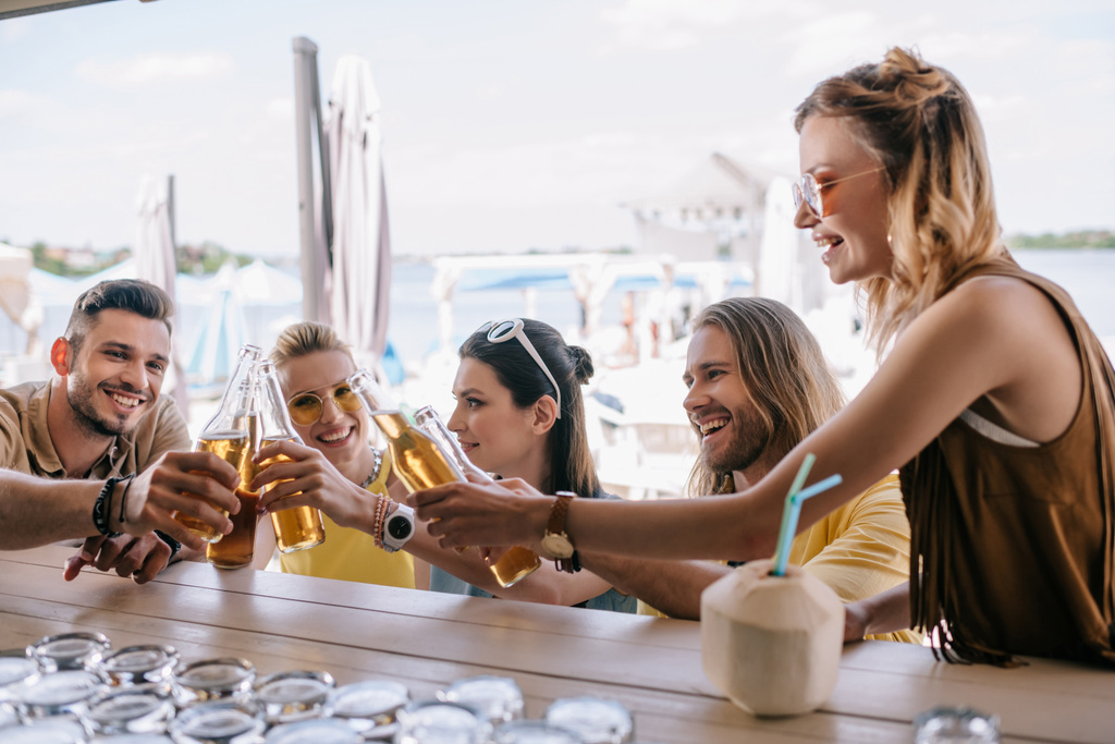 Улыбающиеся юные друзья звонят бутылками пива в пляжном баре
 - Фото, изображение