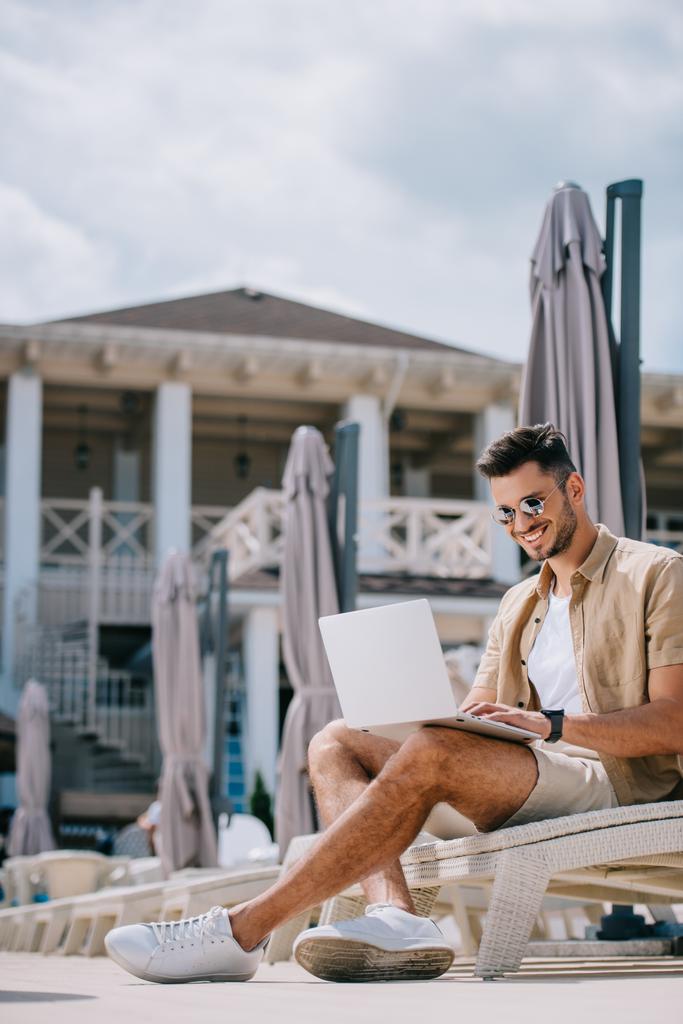 усміхнений молодий чоловік в сонцезахисних окулярах сидить на довгому ланцюжку і використовує ноутбук
 - Фото, зображення
