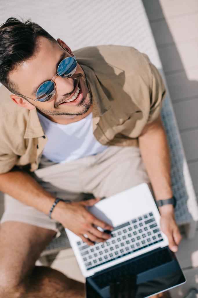 yleisnäkymä nuoresta miehestä aurinkolaseissa kannettavan tietokoneen avulla ja hymyilemällä kameralle
 - Valokuva, kuva
