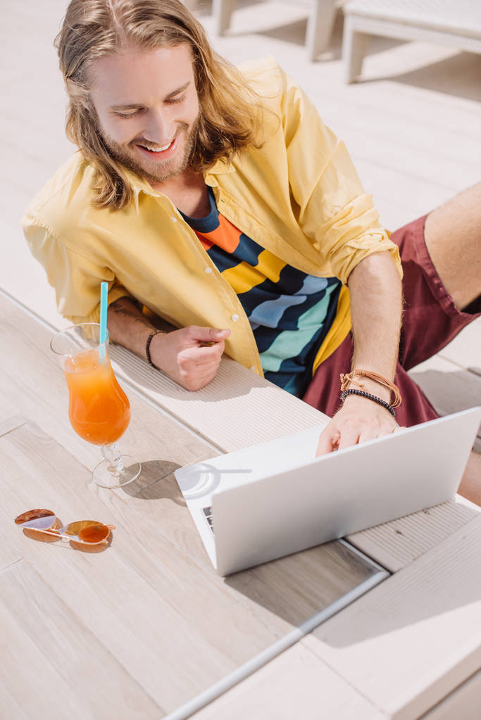 giovane uomo sorridente che utilizza il computer portatile mentre riposa con cocktail a bordo piscina
  - Foto, immagini