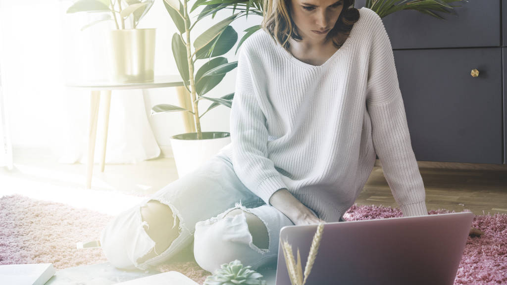junge attraktive Mitarbeiterin arbeitet an einem Laptop, während sie auf rosa Teppich im hellen Wohnzimmer des modernen Hauses sitzt. - Foto, Bild