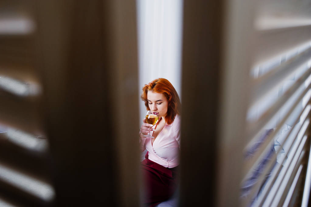 Πανέμορφα κόκκινα μαλλιά κορίτσι στο ροζ μπλούζα και κόκκινη φούστα με ποτήρι κρασί στο χέρι στο κρεβάτι στο δωμάτιο. - Φωτογραφία, εικόνα